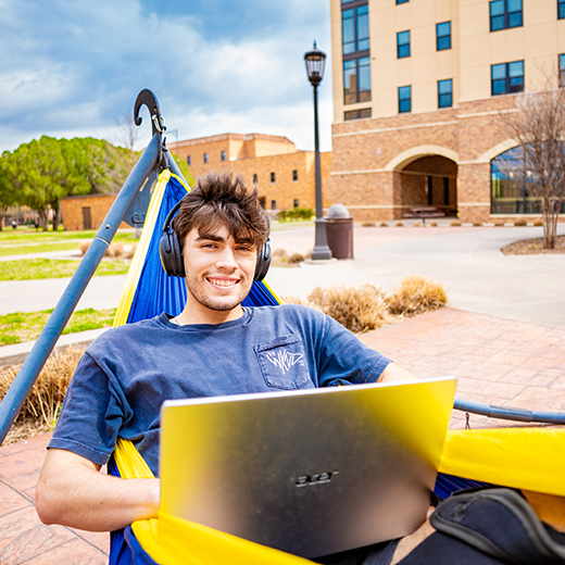 Student studying outside in a hammock, with a Dell laptop, while wearing headphones.