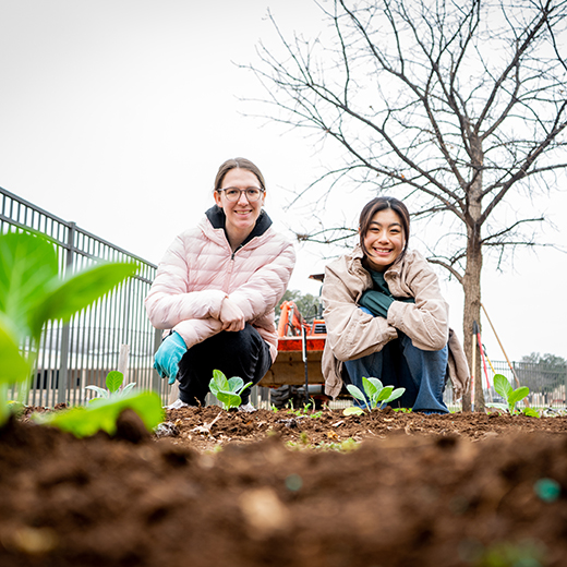 Two smiling honors students pose in a garden bed near blossoming greenery.