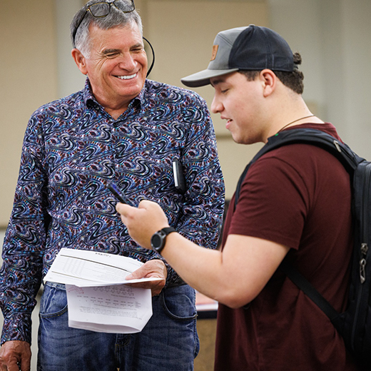 A professor chats with a student who is scrolling on their phone, while talking with the professor.