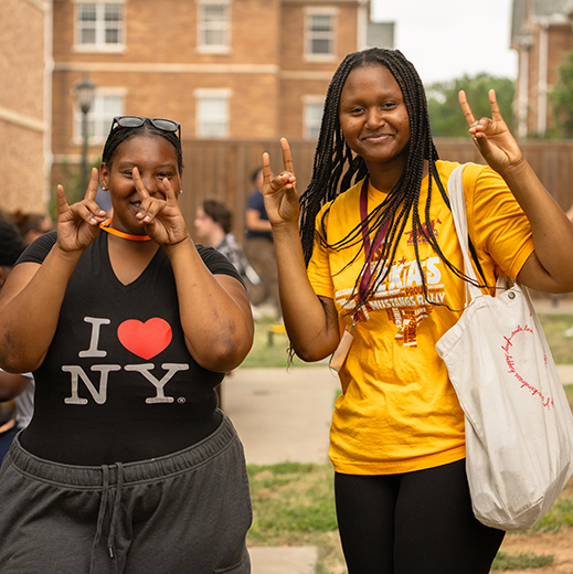 Two students holding up the MSU Texas hand sign while smiling, standing outside of campus housing.