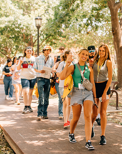 A group of smiling students walk through the quad, while several hold up the Mustangs Hand Sign.