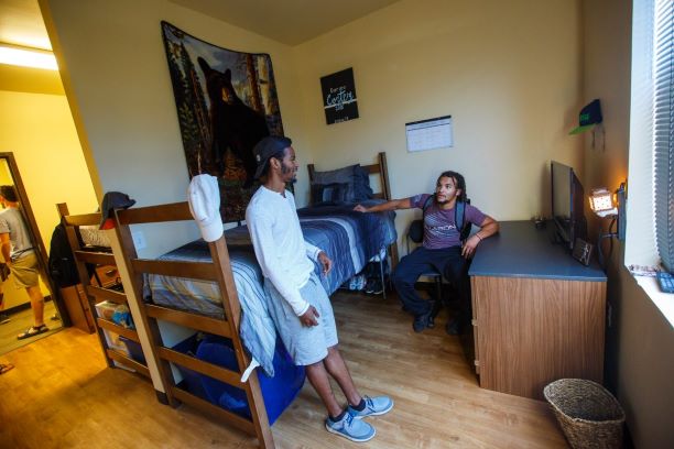 Legacy Hall, room interior, side 'B'. Two students are hanging out in a Legacy Hall room on the window side, side 'B'. The window is along the wall on the right-hand side and has the blinds down, muting the light in the room. Along that same way running towards the back wall is a desk and dresser pushed together to create a large space, a tv sits on the desk and a shelf with a hat is hung on the wall above it. One student is sitting in a rolling chair with one arm leaning against the desk and the other touching the lofted bed on the opposite wall. The second student is leaning against the bed which is raised halfway up. There is a bean bag chair under the bed along with shoe storage. The bed is up against the wall that divides the room in half. Warm light is coming from side 'A' and you can see the end of the other bed positioned along the opposite side of the dividing wall. A few students are standing talking in the open doorway and you can make out part of a dresser on the wall next to the door, but no other part of side 'A' can be seen.