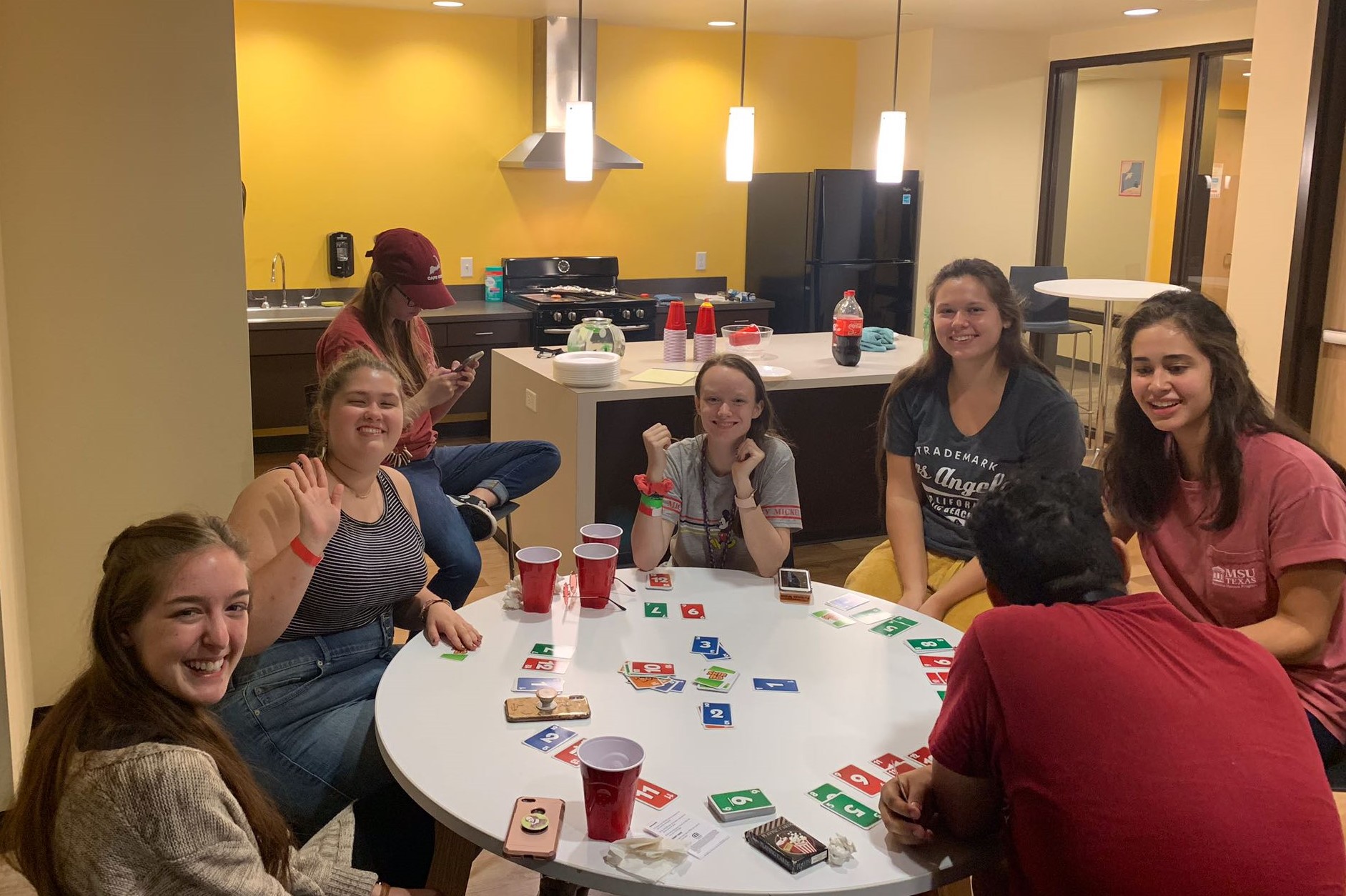 Honors Lounge in Legacy Hall, Interior. Seven Honors students are hanging out in the Legacy Hall Honors Lounge playing Uno. They are seated at a round table. The full-size kitchen can be seen in the background with drinks set up on the kitchen island. A full-size refrigerator, stovetop and oven, exhaust, and large sink can be seen along the far wall. Windows along the right wall near the door reveal part of the Honors hall. 