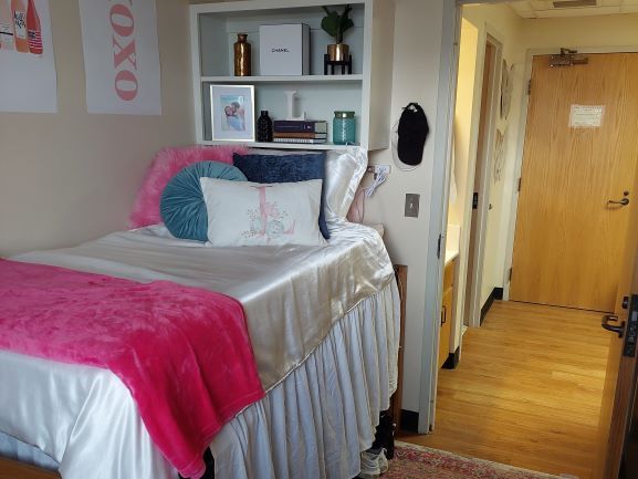 McCullough-Trigg Hall, bedroom interior. A lofted bed sits along the left wall, the mattress is even with the door handle providing extra storage space below the bed. We can see a few pairs of shoes sticking out under the long bed skirt. A built-in double shelf sits above the head of the bed with decorations, books, and pictures. The only other thing along the wall with the shelves is the door to the room which is currently open to reveal the shared living area and the main door to the suite. The vanity can be seen just outside of the bedroom door on the left and a second door leading to the bathroom. A rug sits in the bedroom covering the laminated wood floor seen throughout the suite.