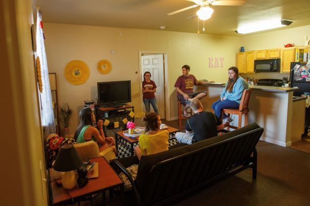 Sundance Two Bedroom Apartment, decorated living room interior. Six students are gathered in the living room area of a Sundance Court two-bedroom apartment. This picture is taken from the corner of the room looking towards the kitchen. From the left, a window can be seen along the wall with curtains One student sits in a chair along the wall, and two more sit on a couch with their backs towards the camera facing a coffee table in the center of the room with a ceiling fan above and tv on a tv stand situated along the far wall. Another student stands in the doorway of one of the bedrooms situated next to the TV. To the right of the living room, a bar area separates the living room from the kitchen and the last two students are sitting on wooden chairs at the bar. Behind them, the top row of cabinets in the kitchen can be seen along with a full-size refrigerator and microwave sitting above a stove. A long fluorescent light is on the ceiling in the kitchen. The carpet in the living room turns into laminated wood floors in the kitchen. 