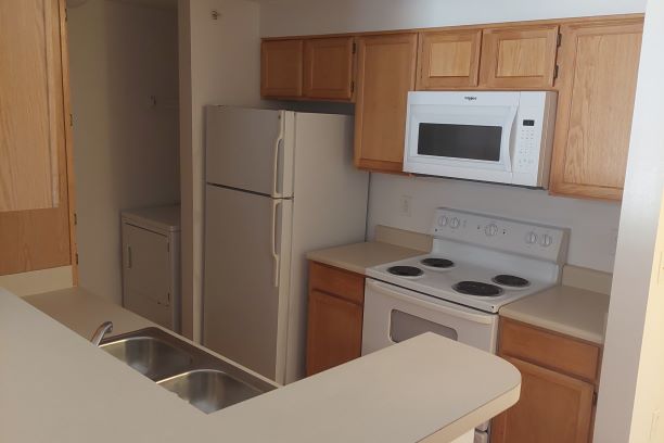 Sunwatcher Kitchen Sunwatcher Village, four-bedroom apartment kitchen, interior, move-in ready. The Sunwatcher Village kitchen is move-in ready meaning that the picture was taken prior to students moving in for the semester. The kitchen sits just off of the entryway. On the right, tan countertops and wooden cabinets line the wall. A stove/oven sits in the middle of the countertop with a microwave mounted above the stove connected to the ventilation system. A full-size refrigerator at the end of the kitchen area. A small wall divides the kitchen from the dryer. Metal shelving can be seen above the dryer. Opposite this wall is another counter space with an elevated bar area, but no wall, so the top section is open to the living room (not seen in this image). A two-compartment sink sits opposite the oven in the center of the countertop and a floor-to-ceiling cupboard sits across from the refrigerator. The washing machine (not in view) is across from the dryer next to the cupboard.