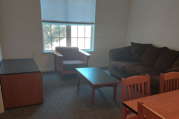 Sunwatcher Village, four-bedroom apartment living room, interior, move-in ready. The Sunwatcher Village living room is move-in ready meaning that the picture was taken prior to students moving in for the semester. This picture was taken standing next to the kitchen looking towards the window on the far wall of the living area. A cushioned chair is positioned in front of the window facing towards a coffee table in the center of the room. To the left, a wooden TV stand sits along the wall. Across from it on the right wall is a large plush couch. A kitchen table can be seen on the right-hand side of the picture against the bar area (not seen in this picture) that divides the kitchen from the living room.