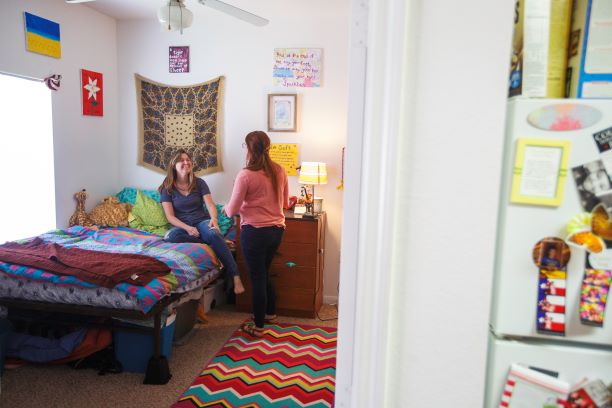 Sunwatcher Village bedroom Sunwatcher Village, two-bedroom apartment, interior. Two students are talking in one of the bedrooms of Sunwatcher Village's two-bedroom/ two-bathroom apartments. The picture is taken just outside of the doorway of the bedroom and you can see part of the refrigerator on the right-hand side of the image. Through the doorway, you can see a full-size bed elevated off the ground allowing for storage underneath. It is positioned against the far corner of the room next to a large window. The dresser has been positioned next to the bed providing a space for a lamp and various items. There is a ceiling fan in the center of the room and a bright rug on the carpeted floor. The right side of the room is not seen due to the door being partially open.
