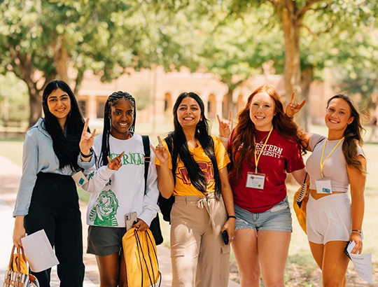 Five students walking through the quad stop to smile and hold up the Mustangs hand sign.