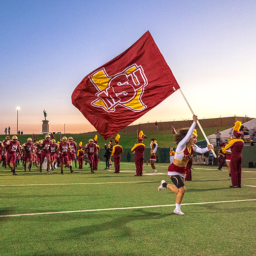 An MSU Texas cheerleader runs with the MSU Texas flag as the Golden Thunder Marching Band plays and the MSU Texas Mustang football players run onto the field at Memorial Stadium.